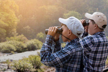 Asian boys wearing t-shirt holding a binoculars sitting on river bank in local national park to observe fish in the river and birds on tree branches and on sky.の写真素材