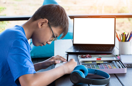 asian boy spends his free times with drawing, coloring, painting and sketching pictures in front of labtop on table in the middle of living room outside his house.の写真素材