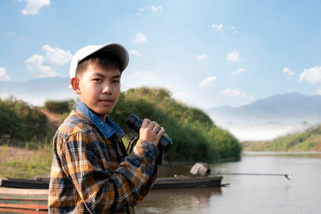 Asian boy in plaid shirt and cap, sitting and holding binoculars on bow of a boat parked beside a river to observe birds and fish swimming in the river during his summer holidays.の写真素材