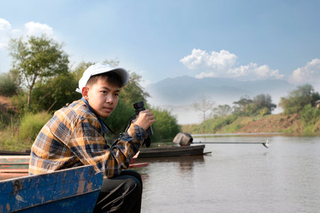 Asian boy in plaid shirt and cap, sitting and holding binoculars on bow of a boat parked beside a river to observe birds and fish swimming in the river during his summer holidays.の写真素材