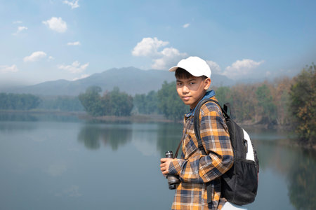 Asian boy in plaid shirt wears cap and has a backpack, holding a binoculars, standing on ridge...