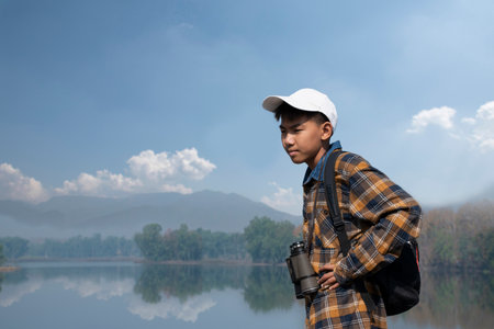 Asian boy in plaid shirt wears cap and has a backpack, holding a binoculars, standing on ridge reservoir in local national park to observe birds and to watch fish in reservoir.の写真素材