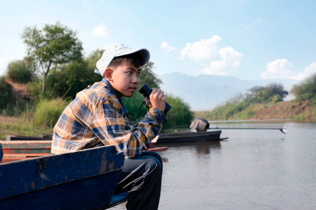 Asian boy in plaid shirt and cap, sitting and holding binoculars on bow of a boat parked beside a river to observe birds and fish swimming in the river during his summer holidays.の写真素材