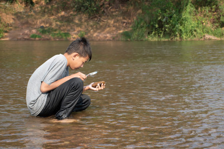 Asian boy sitting on the bank of the river and holding a magnifying glass to see the rock and underwater creature.の写真素材