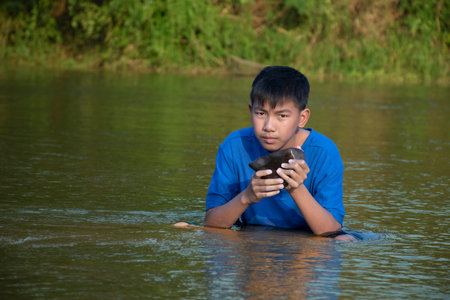 Asian boy in blue t-shirt is spending his freetimes by diving, swimming, throwing rocks and catching fish in the river happily, hobby and happiness of children concept, in motion.の写真素材