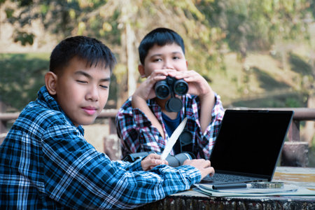 Two young boys using a laptop and binoculars in the parkの写真素材