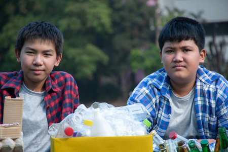 Asian boys in plaid shirt sitting on playground of school with various boxes of separated garbabes...