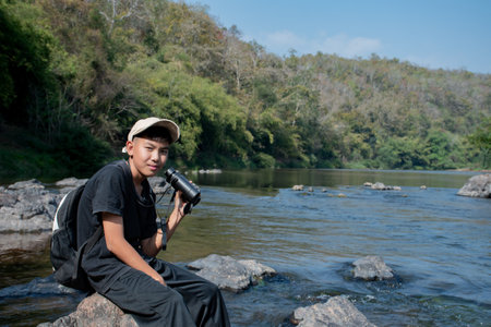 Young asian photographer sitting on a rock with binoculars in the riverの写真素材