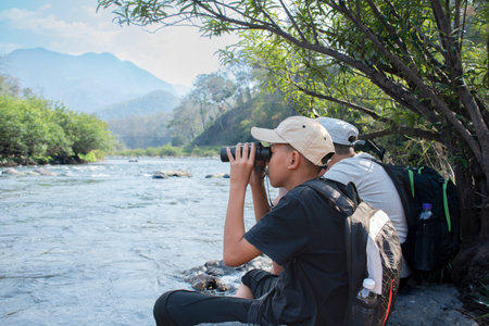 Young asian man sitting on the bank of a mountain river and looking through binocularsの写真素材