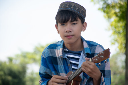 Asian cute muslim or islamic boy sitting and playing ukulele under the tree in local community park, recreational activity and hobby of all children around the world concept.の写真素材