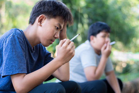 Asian boy is trying to smoke with his friend in the public park, concept for bad behavior of young teenboys around the world.の写真素材