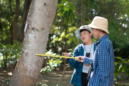 Young asian schoolboy measuring a size of tree trunk with a measuring tape and recording information...