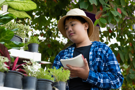 Asian cute boy holds pen and notebook near various potted houseplants and recording and learning the growing of houseplants, soft and selective focus.の写真素材