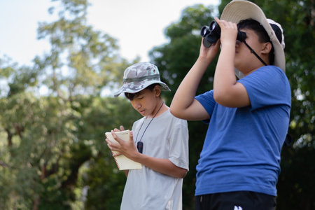 Two Asian boys exploring nature, one using binoculars and the other writing notes. Outdoor learning, curiosity, and childhood adventure in a green, forest-like environment.の写真素材