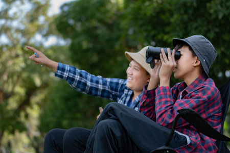 Two Asian boys exploring nature, one using binoculars and the other writing notes. Outdoor learning, curiosity, and childhood adventure in a green, forest-like environment.の写真素材
