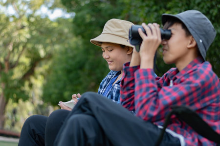 Two Asian boys exploring nature, one using binoculars and the other writing notes. Outdoor learning, curiosity, and childhood adventure in a green, forest-like environment.の写真素材