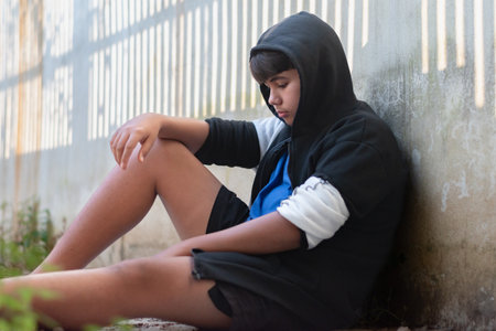 Asian boy in a black hoodie sits with his knees pressed against a dirty wall.の写真素材