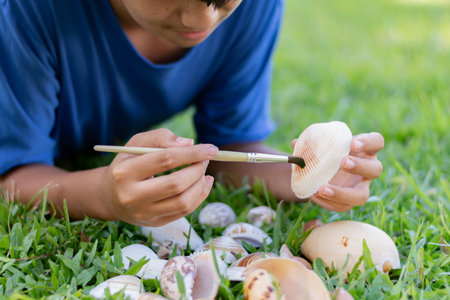 Boy painting seashells with brush on green grass in the parkの写真素材