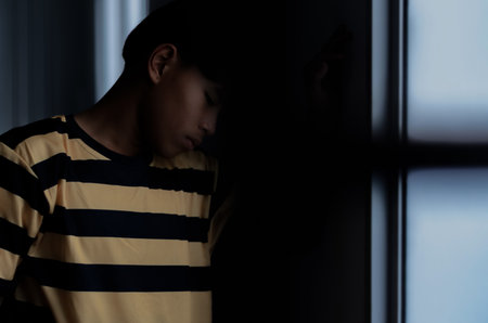 Teenage boy with depression looking through window in dark room, closeupの写真素材