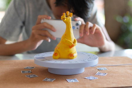 Young Asian man capturing small giraffe piggy bank object on turntable to make 3D  video with mobile...