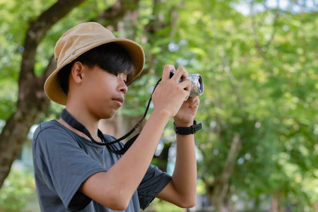 Side view of a concentrated young boy in a bucket hat taking a picture outdoors with a digital camera against a bright green, natural background.の写真素材