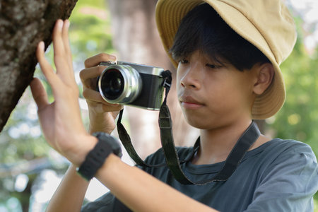 A young photographer in a bucket hat taking a close-up shot of tree bark with a camera, showing interest in nature and texture.の写真素材