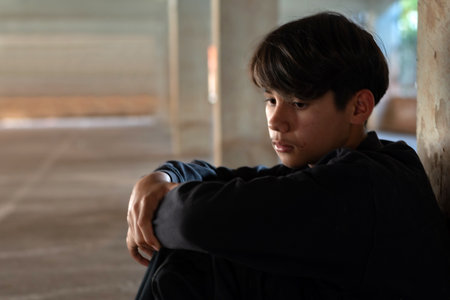 Close-up portrait of a sad, lonely teen boy in dark clothes sitting in a dark, sheltered area,...