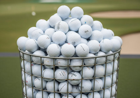 A close-up of a wire basket filled to the brim with numerous white golf balls, set against a blurred background of a golf course.の素材