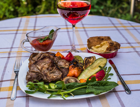 Delicious juicy grilled steak, vegetables and mushrooms on the plate, surrounded by tomatoes, fresh herbs and red barbecue sauce. Glass of red wine and lavash.の写真素材