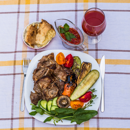 Delicious juicy grilled steak, vegetables and mushrooms on the plate, surrounded by tomatoes, fresh herbs and red barbecue sauce. Glass of tomato juice and lavash.の写真素材