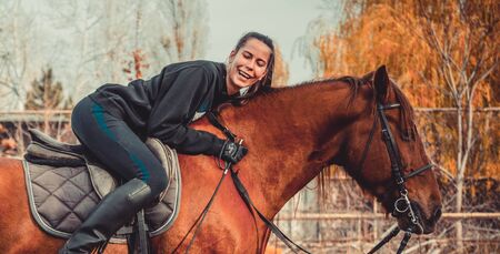 Young beautiful brunette girl rides a horse on a warm and sunny autumn day. Portrait of a pretty young woman on the horse, wearing tall boots and gloves.の写真素材