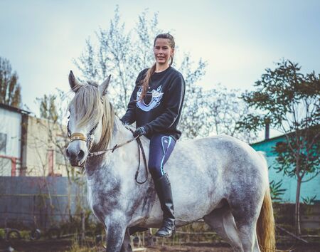 Young beautiful brunette girl rides a horse bareback with hackamore. Warm and sunny autumn day. Portrait of a pretty young woman on the horse, wearing tall boots and gloves.の写真素材