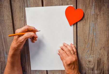 Elderly woman hand going to write a letter, red heart on rustic wooden table as a background, sun rays. View from above, copy space. Mother or Woman, Valentine day, human emotions, love and compassion, warmth, senior concept.の写真素材