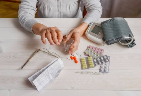 Senior hands with medicine bottle putting drops in a glass, pills, tonometer, thermometer on a table. Woman wrinkled hands, colorful tablets, blood pressure cuff. Elderly health care, old age.の写真素材