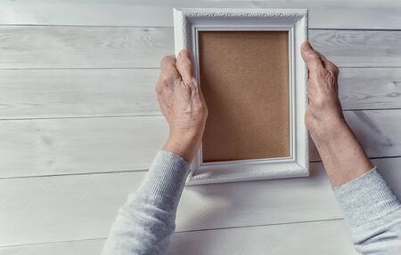 Senior wrinkled hands holding white photo frame. Elderly woman looking at the picture, light wodden...