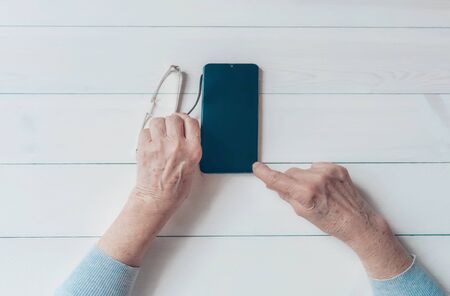 Old wrinkled hands holding mobile smart phone. Elderly woman using digital phone showing something on display, light table. Template, copy space mockup. Teaching, technologies for seniors concept.の写真素材