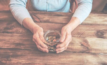 Old wrinkled hands holding jar with coins, wooden background. Saving money for future plan and retirement fund, pension, poorness, need concept.の写真素材
