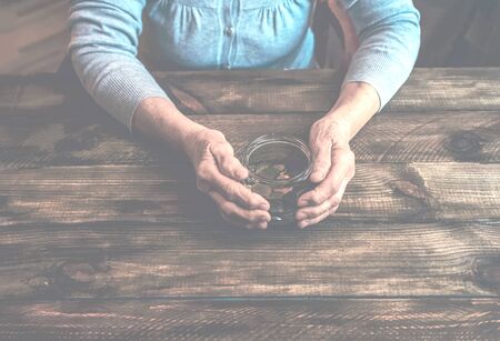 Old wrinkled hands holding jar with coins, wooden background. Saving money for future plan and retirement fund, pension, poorness, need concept.の写真素材