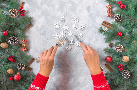 Christmas gray grunge concrete background, fir twigs, berries, snowflakes, baubles, top view. Woman hands in red pullover, fork and knife, white snowflakes. New Year table setting menu decoration.の写真素材