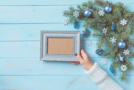 Christmas light blue background, woman hand in white pullover holds frame for wishes. Fir branches, blue silver baubles, stars, hearts, snowflakes. New Year festive decoration, top view, copy space.の写真素材