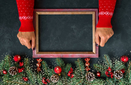 Christmas dark grunge horizontal background, top view. Fir twigs, red golden berries, baubles, New Year decoration. Woman hands in patterned red sweater hold frame for wishes, copy-space.の写真素材