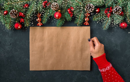 Christmas dark grunge horizontal background, top view. Fir twigs, red golden berries, baubles, New Year decoration. Woman hand in patterned red sweater holds aged paper, copy-space.の写真素材