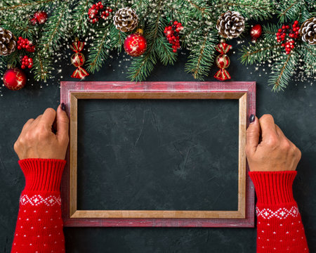 Christmas dark grunge horizontal background, top view. Fir twigs, red golden berries, baubles, New Year decoration. Woman hands in patterned red sweater hold frame for wishes, copy-space.の写真素材