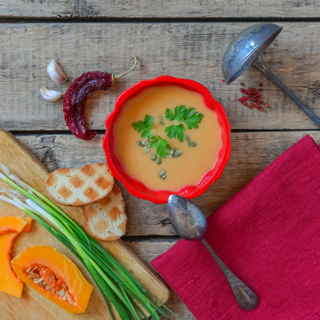Pumpkin soup on wooden table, linen cloth, vintage cutlery. Autumn cream soup, seeds and parsley. Sliced herbs, pumpkin on cutting board, cooking process. Thanksgiving dinner, flat lay, top view.の写真素材