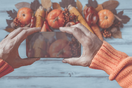 Autumn background, fallen leaves, fruits, vegetables, rustic wooden table. Hands taking photo with mobile phone. Seasonal set, pumpkins, corn cobs, wheat. Thanksgiving food, healthy, fresh, top view.の写真素材