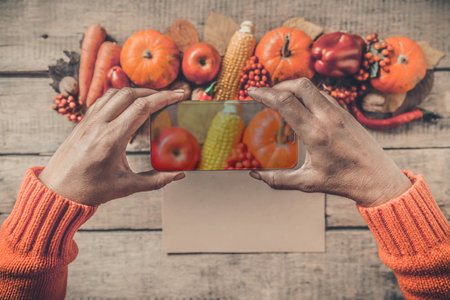 Autumn background, fallen leaves, fruits, vegetables, rustic wooden table. Hands taking photo with mobile phone. Seasonal set, pumpkins, corn cobs, wheat. Thanksgiving food, healthy, fresh, top view.の写真素材