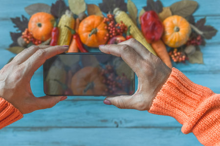 Autumn background, fallen leaves, fruits, vegetables, rustic wooden table. Hands taking photo with mobile phone. Seasonal set, pumpkins, corn cobs, wheat. Thanksgiving food, healthy, fresh, top view.の写真素材