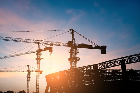 Building site with high-rise block under construction in an urban environment dominated by a large industrial crane silhouetted against a cloudy blue skyの写真素材