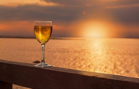 A glass with a cool drink standing on a wooden balustrade in the foreground of the seaside where you can see the sun down with golden light.の写真素材