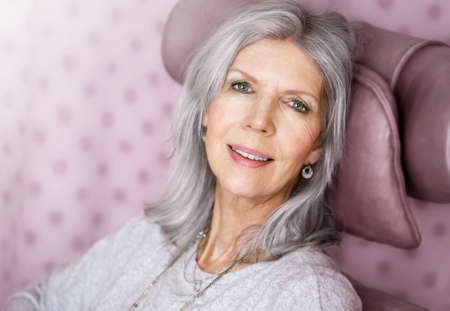 Happy mature woman sitting to lean her head on backrest in front of an dusky pink background, to...
